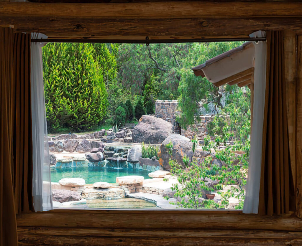 A serene pool view framed by a rustic wooden window, surrounded by lush greenery.