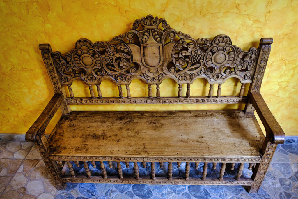 An ornately carved wooden bench against a textured yellow wall exudes rustic elegance, blending artisanship with inviting warmth.