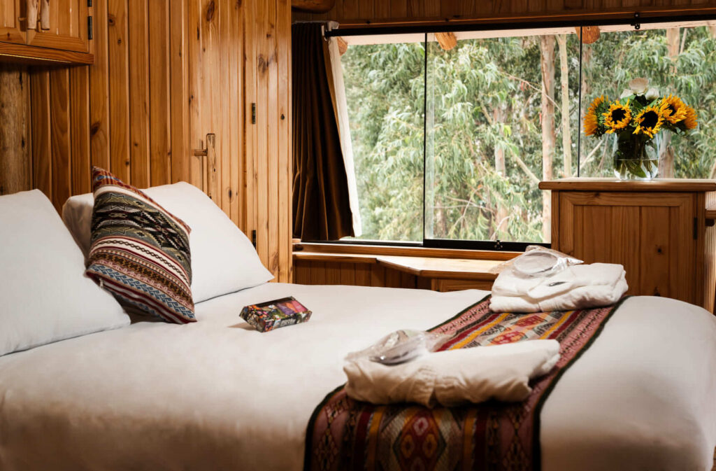 The Sunbeam Suite at Ayahuasca Lux presents a harmonious blend of comfort and natural elegance, with its plush white bedding contrasting the warm wooden interior and large windows framing the tranquil forest views.