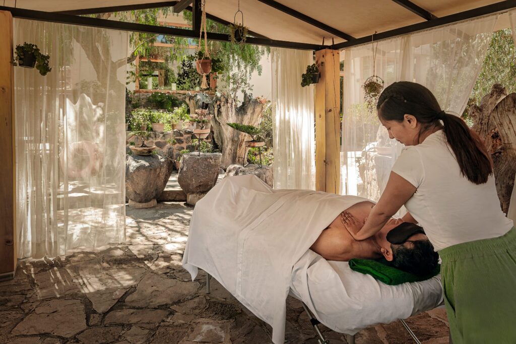 a peaceful spa setting where a person is receiving a massage. The environment looks relaxing, with natural light and greenery surrounding the area, enhancing the soothing experience of the spa treatment