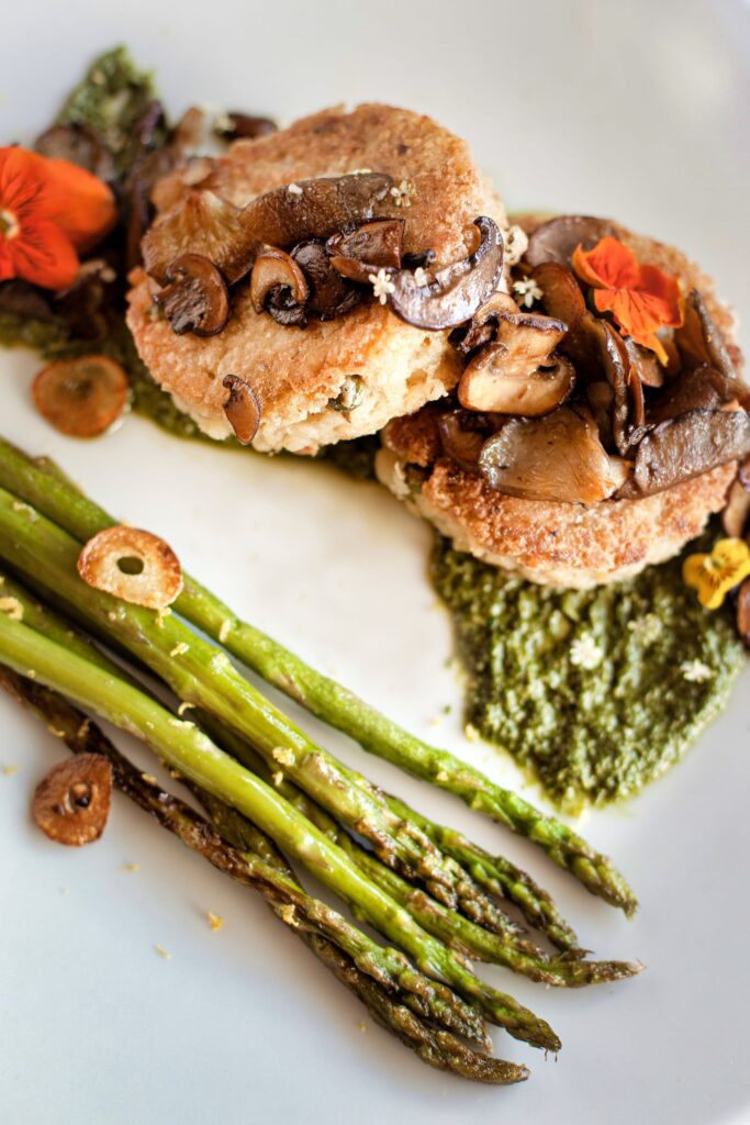 a gourmet dish with sautéed mushrooms atop a golden-brown crusted patty, served with grilled asparagus on a bed of vibrant green sauce, garnished with a delicate edible flower.