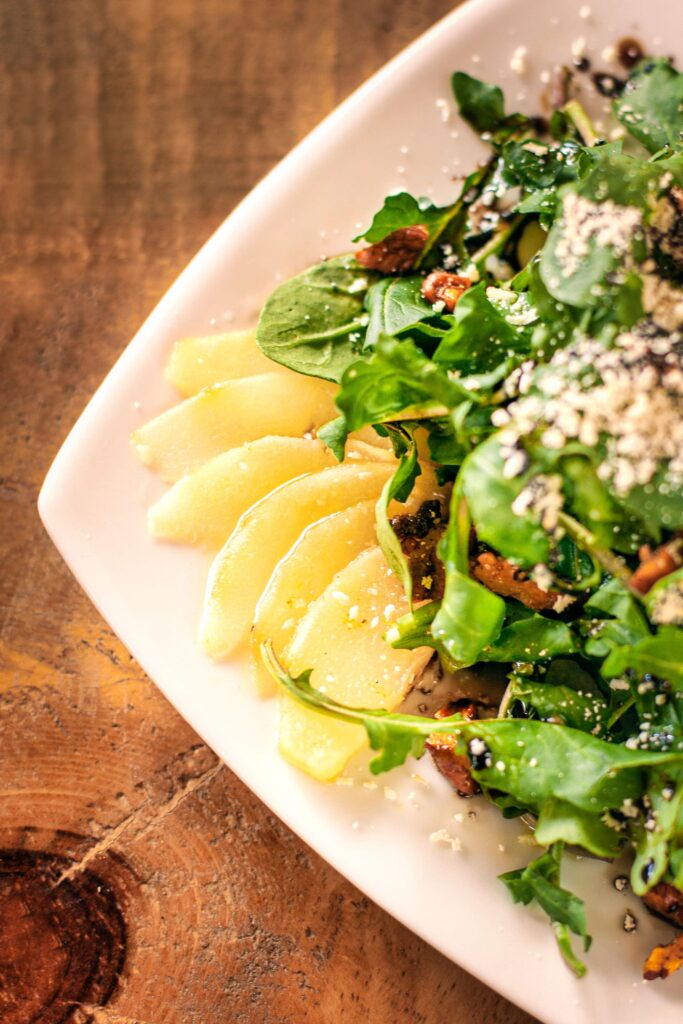 The image features a fresh, gourmet salad with crisp greens, perhaps baby spinach or arugula, topped with toasted nuts and a generous sprinkle of crumbled cheese. Thinly sliced pears fan out on one side, adding a sweet and juicy element